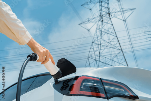 Closeup woman recharge EV electric car battery at charging station connected to electrical power grid tower on sky background as electrical industry for eco friendly vehicle utilization. Expedient photo