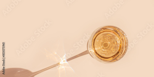 One wineglass with white wine sparkling at sunlight, trend peach beige background with dark shadow and reflection, summer aesthetic minimal banner. Shiny Alcohol drink, above, neutral pastel color