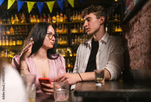 Guy in wrinkled shirt loses smartphone and asks girl to call number at noisy party. Girl helps guy find mobile phone in bar at night