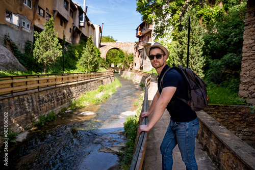 A young man explores new destinations by traveling through North Macedonia.He is standing by the river and looking at the camera.