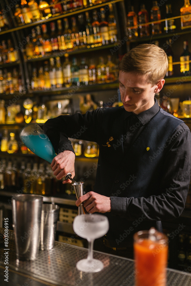 Barman pours blue syrup for cocktails into jigger to measure. Young worker in uniform makes cocktail Blue Lagoon for elegant guests of bar