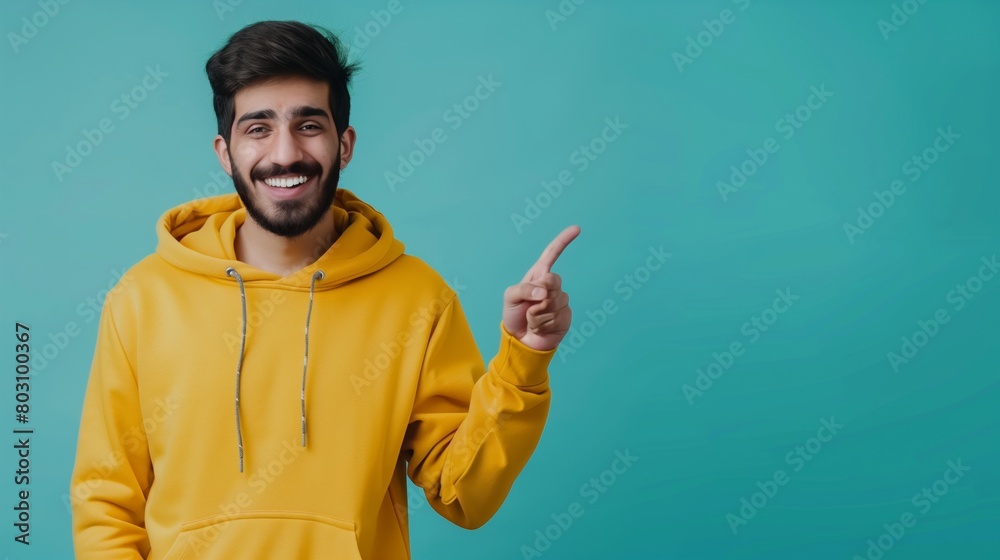 Cheerful young man in a yellow hoodie pointing to the side on a teal background. Cheerful young man in a yellow hoodie pointing to the side on a teal background.