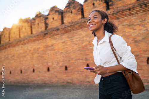 black girl holding phone ancient area finding location on holiday. female teenage tourist holding baggage looking for chiangmai gateway view. african american girl searching for landmark brick wall