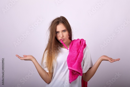 A young woman with a thoughtful expression stands with a toothbrush in her mouth and a towel on her shoulder. She raises her hands in the air, looking at the camera with a questioning expression.