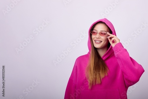 A stylish young woman wearing a pink hoodie and sunglasses poses for a photo. She has a happy expression on her face and is looking away from the camera. The background is a solid white color.