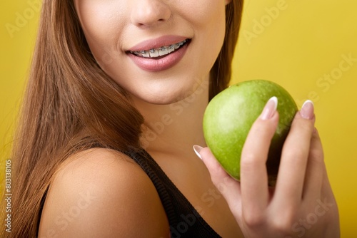 A young woman with braces on her teeth is holding a green apple in front of her mouth. She has long brown hair and is wearing a black tank top. The background is a bright yellow color.