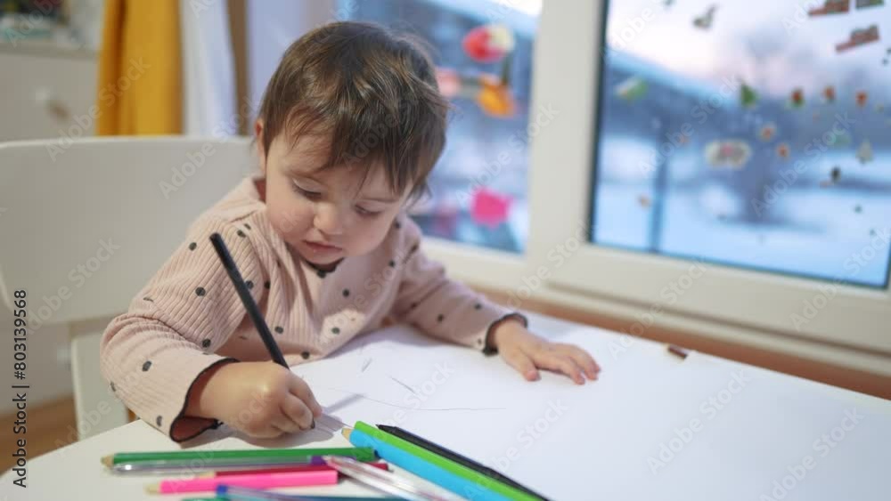 baby girl draws while sitting at a table by the window at home. happy family kid concept. baby daughter learns to draw with pencils dream on a sheet of paper indoors. development of fine motor skills baby girl draws while sitting at a table by the window at home. happy family kid concept. baby daughter learns to draw with pencils dream on a sheet of paper indoors. development of fine motor skills