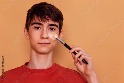 A male teenager puts a moisturizing mask or cream on his face. Male teen in casual t-shirt is smiling and holding a brush. Hygiene and skin care in adolescence.
