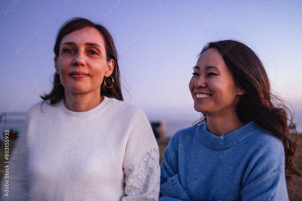Two smiling multiethnic friends in sweaters enjoy nature in the evening. Young cheerful women of different races travel and have fun together outdoors.