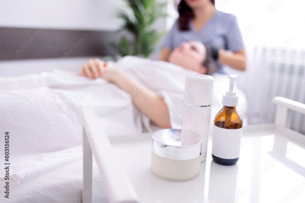Mock-ups of beauty cosmetology products on table in cosmetologist office in spa salon or clinic. Dermatologist and her client in background are blurred. Mock-ups of beauty cosmetology products on table in cosmetologist office in spa salon or clinic. Dermatologist and her client in background are blurred.