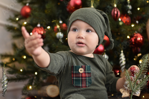 Baby 1 year old sitting in chair with Christmas tree and lights on background in room. Merry Christmas. Holiday season