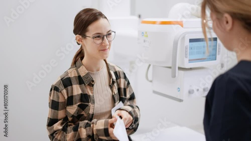 A happy brunette girl in round glasses in a plaid shirt receives the results of her fluorography in pictures in a special package from a girl doctor in the fluorography room in a modern clinic photo