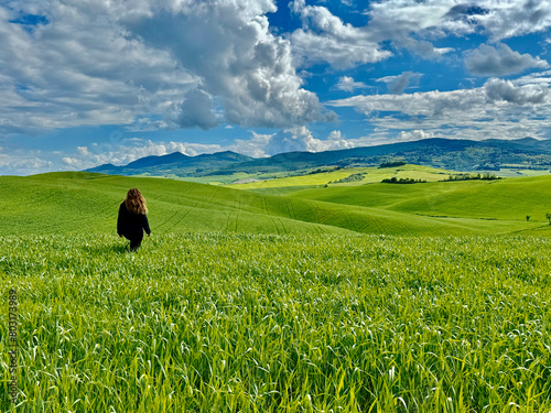 A woman walks through lush green fields under a clear blue sky, Tuscany hills, Italy.