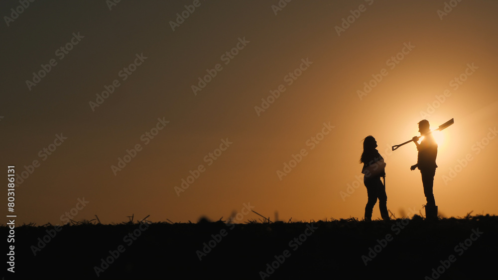 Two farmers stand in a field at sunset. Man holding a shovel, talking to a woman
