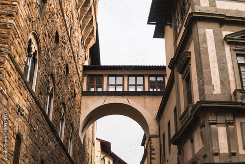 Medieval charm captured on Via della Ninna: Palazzo Vecchio's side adorned with medieval windows connected to an arch bridging to the neighboring building under the cloudy sky.