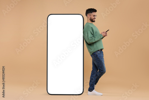 Man with mobile phone standing near huge device with empty screen on dark beige background. Mockup for design