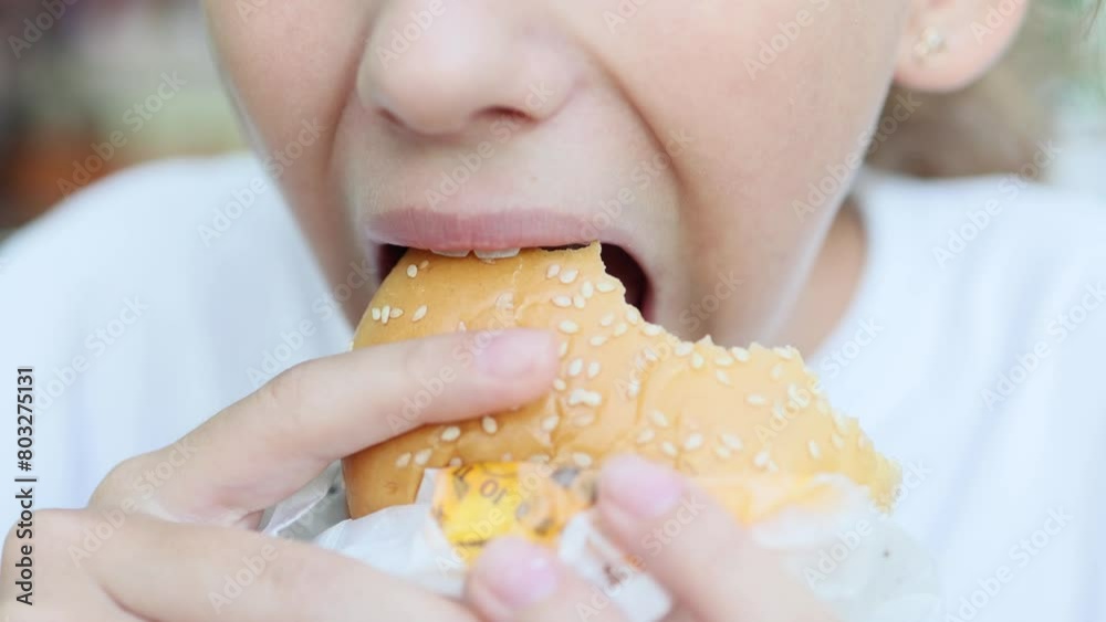 A cute blonde teenage girl eats a delicious burger with an appetite at a fast food restaurant. The harm of fast food for children. Close-up. A cute blonde teenage girl eats a delicious burger with an appetite at a fast food restaurant. The harm of fast food for children. Close-up.