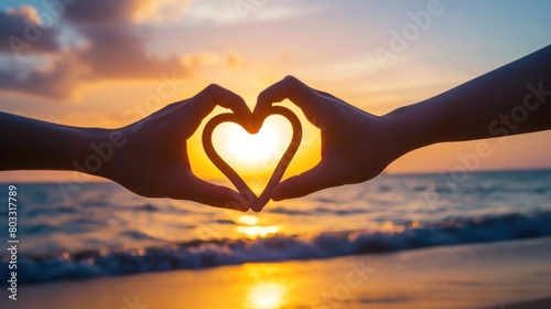 A heart shape made in the style of two hands against the backdrop of sunset on beach