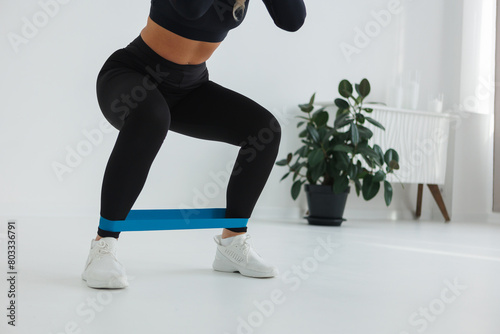 A woman is squatting with a blue resistance band, wearing sportswear. Her knee and thigh are engaged while her human leg is at a right angle. Nearby is a flowerpot with a plant