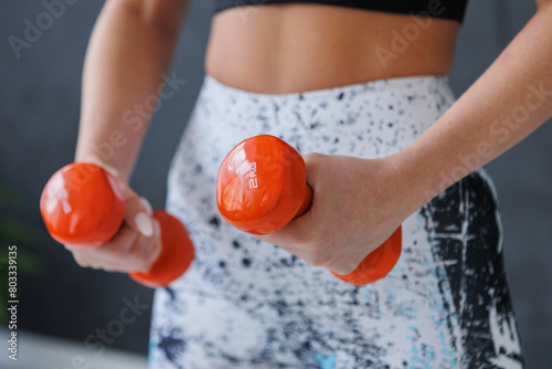 A woman is holding two orange dumbbells in her hands, her fingers wrapped around the handles as her thumb grips the weight. She stands with a straight posture, her waist and elbows in alignment