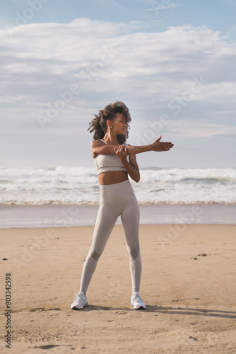 Fit black woman doing arm and shoulder stretching exercise at the beach. Healthy and fitness lifestyle. Outdoor workout.