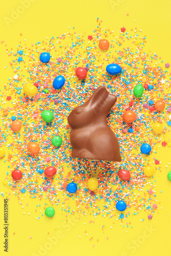 Chocolate bunny and colored sugar sprinkles on a yellow background, top view. Easter composition. Easter chocolate bunny and candy. Festive sweet treat. Chocolate figurine. Sweets