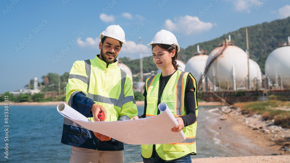 Assured engineer wearing a safety helmet and high-visibility vest holds blueprints, with industrial tanks in the background.
