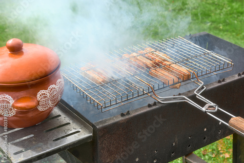Appetizing meat sausages are fried on a smoky grill. A grill for cooking meat and a clay pot for ready-made pieces.