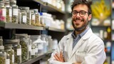 A cheerful male pharmacist with eyeglasses stands confidently in an herbal medicine shop surrounded by jars