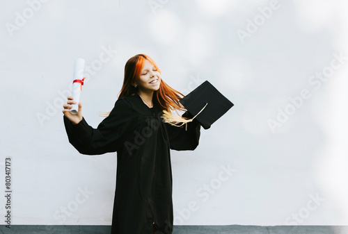 teenage girl in clothes of a graduate coat and cap celebrates high school or  junior year graduation on background of white wall with shadows and with diploma in hands, education and no school concept
