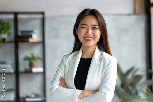 Portrait of a radiant young asian woman in a white blazer, standing arms crossed in a modern office setting, embodying professionalism and confidence with a beaming smile