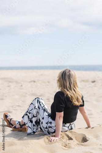 Back of blonde woman in black top, black and white graphic print pants and black sandals sitting in the sand looking at the ocean