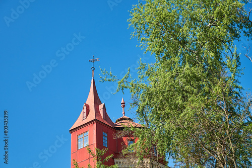 A photorealistic depiction of a quaint red tower with a weathervane perched atop its steeple. The tower stands amidst a lush green landscape, bathed in the warm hues of the setting sun.