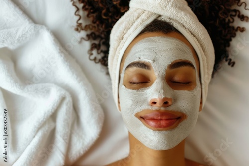 Young woman enjoys a moment of relaxation with a rejuvenating facial mask, lying on a soft white blanket with eyes closed and a gentle smile