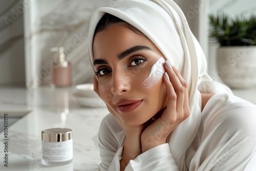 Elegant woman in a headscarf gently applies moisturizing cream on her cheek, showcasing a daily skincare routine in a bright and modern bathroom setting
