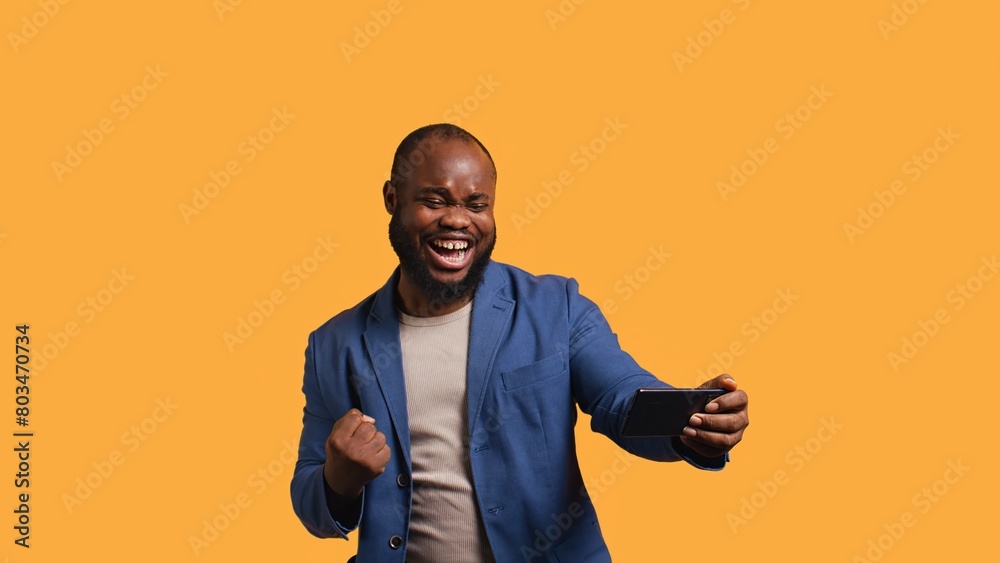 Happy man playing videogames on smartphone, celebrating after defeating foes, clenching hand gesture. Passionate BIPOC player thrilled about winning game on phone, studio background, camera B