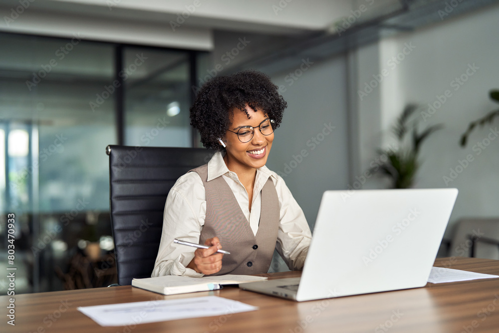 Happy professional young African female hr manager, smiling business woman leader in office wearing earbuds looking at laptop computer talking having hybrid work meeting or remote job interview. Happy professional young African female hr manager, smiling business woman leader in office wearing earbuds looking at laptop computer talking having hybrid work meeting or remote job interview.