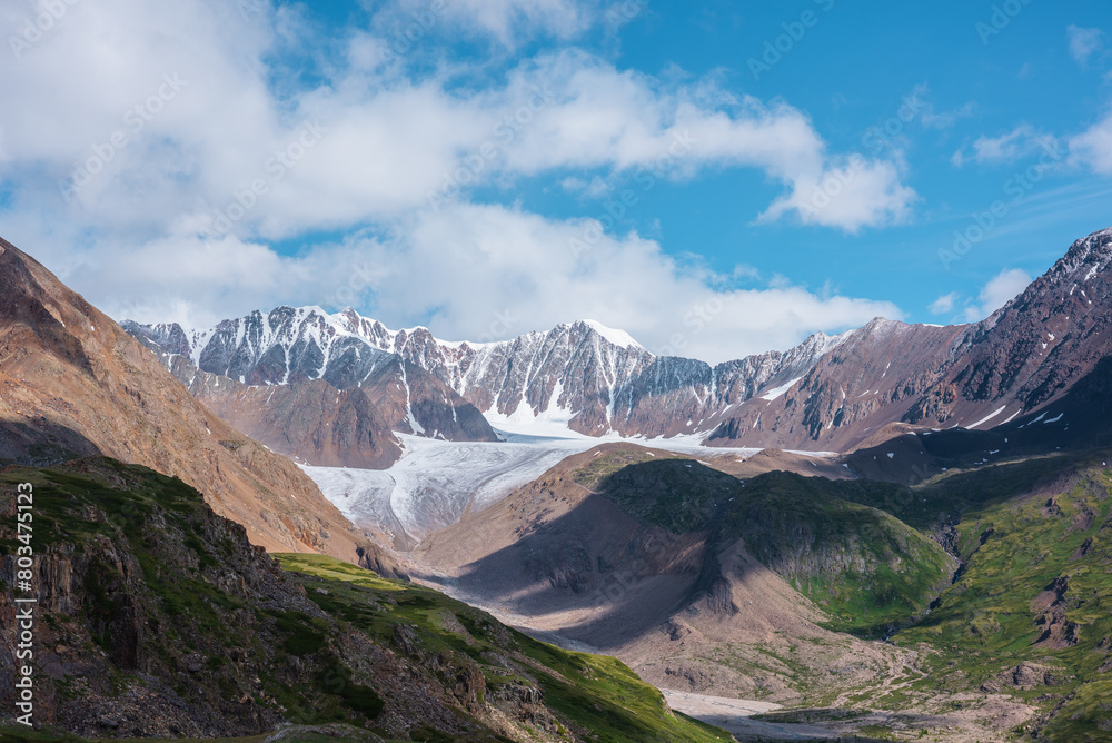 Scenic landscape with glacier tongue among rocky ridges and sharp snow-capped mountain range in sunlight under clouds in blue sky. Alpine valley with green hills and rocks against ice and sheer crags.