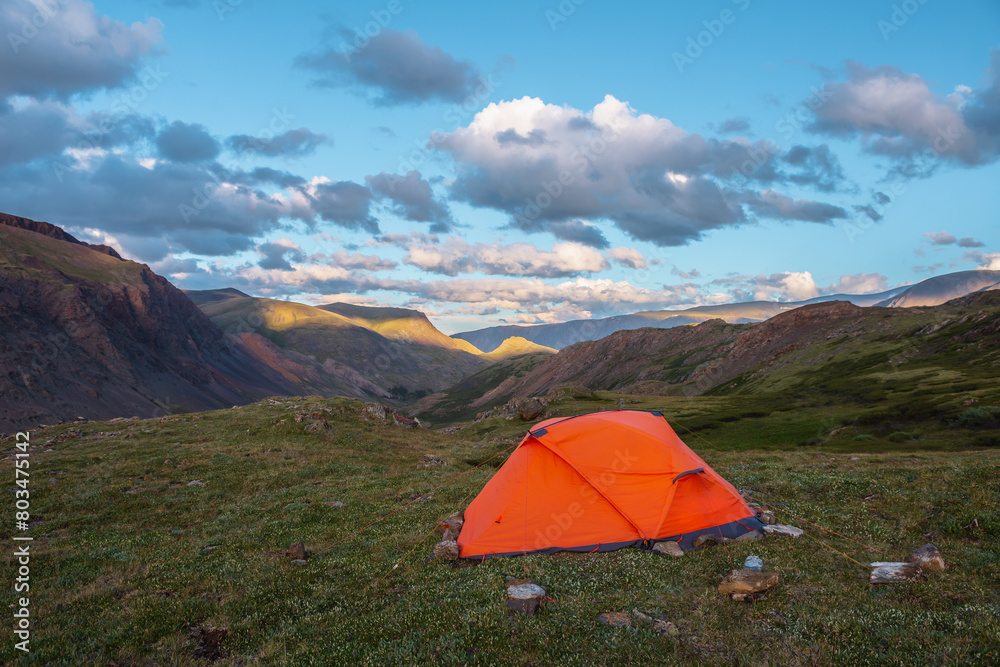 Vivid orange tent on grassy hill with view to mountain tops illuminated by sunset light. Rocks silhouettes. Shadows of clouds on rocky ridge in sunrise colors. Red tent and sunset tones in blue sky.