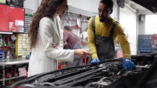 Young auto mechanic explaining to his female customer what needs to be fixed in her car, while his colleague working in the background photo