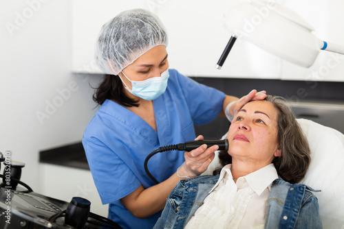 Obraz na plátně Elderly woman getting facial electroporation procedure from experienced cosmetologist in aesthetic medicine office