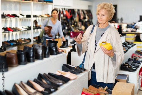 Elderly woman comparing moccasins in different colours while standing in shoeshop. Asian woman shopping in background.