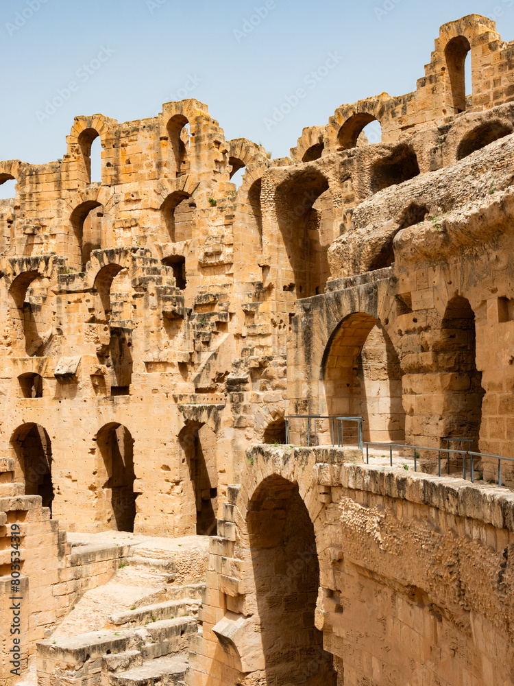 Scenic view of time weathered honey-colored limestone walls of antique Roman amphitheater in Tunisian city of El Djem, with surviving arched openings basking in warm sunlight..