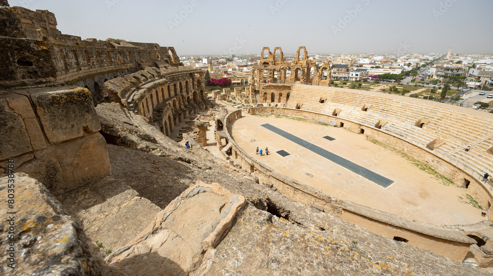Breathtaking view of sun-drenched ancient Roman amphitheater in Tunisian city of El Djem with restored arena, stone seating tiers and crumbling walls with archways against backdrop on modern cityscape