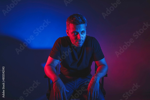 A man in a black shirt sits in a chair in front of a blue background