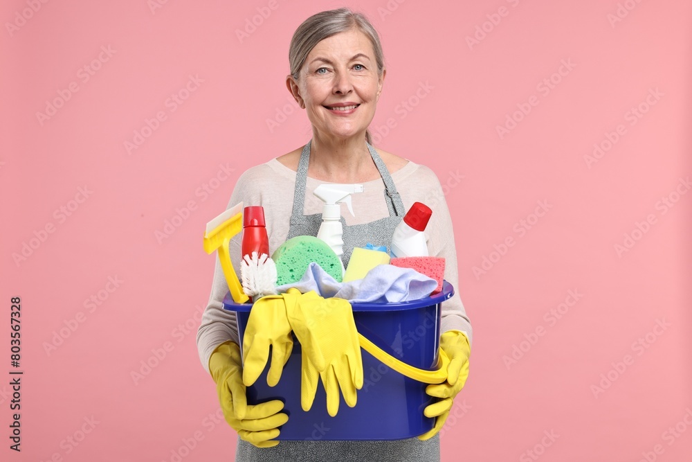 Happy housewife holding bucket with cleaning supplies on pink background Happy housewife holding bucket with cleaning supplies on pink background