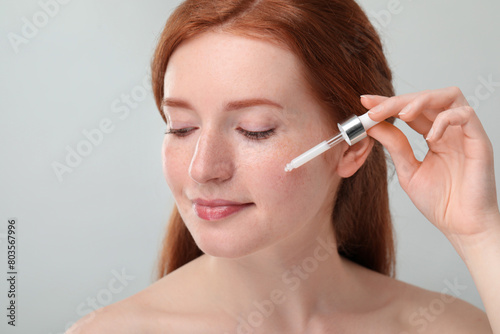 Beautiful woman with freckles applying cosmetic serum onto her face against grey background  closeup