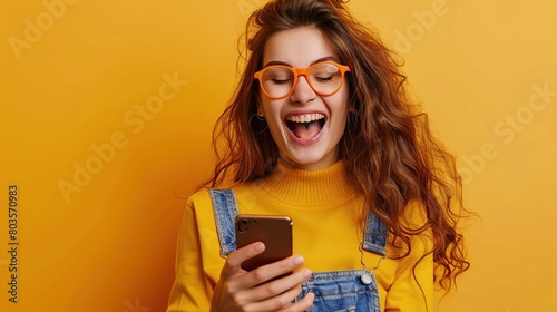 A surprised young lady using a cell phone with a positive expression, wearing casual clothes, glasses and standing isolated on an orange background. Happy and adorable woman, rejoicing in success photo