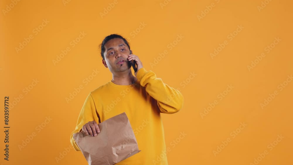 Indian man holding thermal backpack answering phone, fulfilling clients orders, isolated over studio background. Food delivery service employee talking with customer during telephone call, camera B Indian man holding thermal backpack answering phone, fulfilling clients orders, isolated over studio background. Food delivery service employee talking with customer during telephone call, camera B