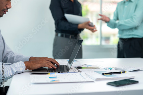 A man is typing on a laptop in front of a table with papers and a cell phone. The scene suggests a busy work environment where the man is working on a project or report